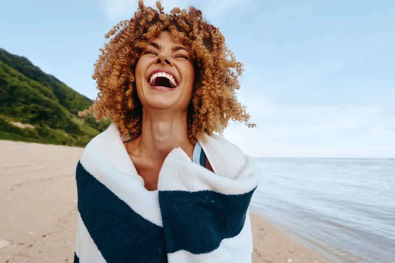 Smiling woman on a sunny beach wrapped in a striped towel, enjoying the breeze by the ocean. Open sky, warm lighting, and a carefree mood evoke summer leisure.
