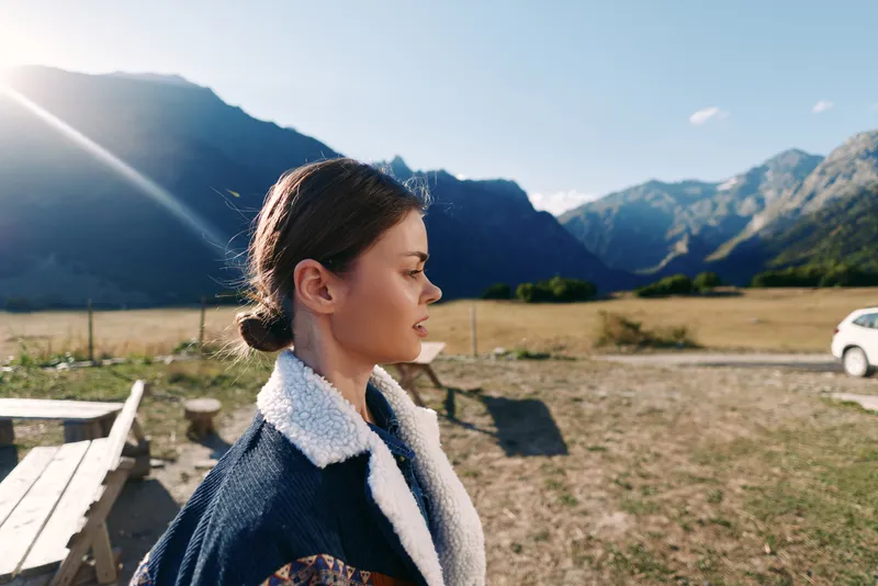 Woman profile portrait in mountains meadow with parked car and picnic table, sunlight flare and warm outdoor scenery. Young traveler in jacket enjoying nature and calm landscape