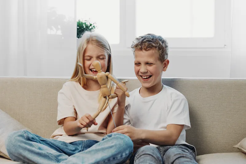 Two happy children sitting on a sofa playing with a wooden artist mannequin, smiling and enjoying a bright cozy home environment, concept of childhood and creativity.