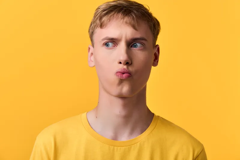 Pouty young man wearing a yellow shirt against a bright yellow backdrop, making a playful kissy face for a lighthearted fashion and expressive mood concept.