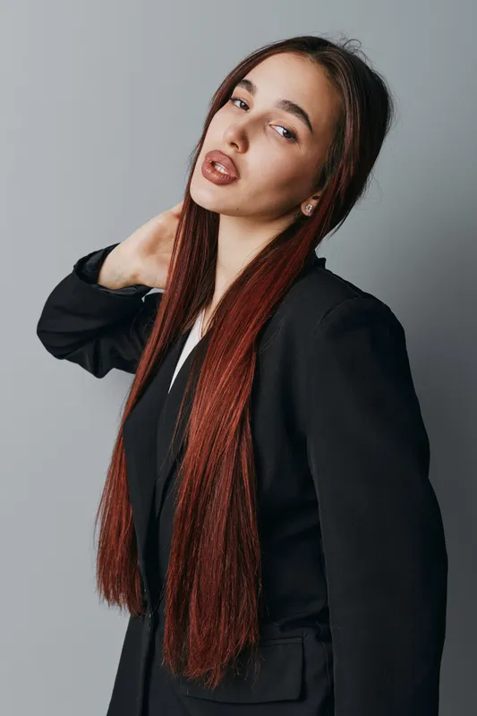 Young woman with long straight hair wearing black blazer posing with hand on neck against gray background. Portrait of confident female model with natural makeup and earrings.
