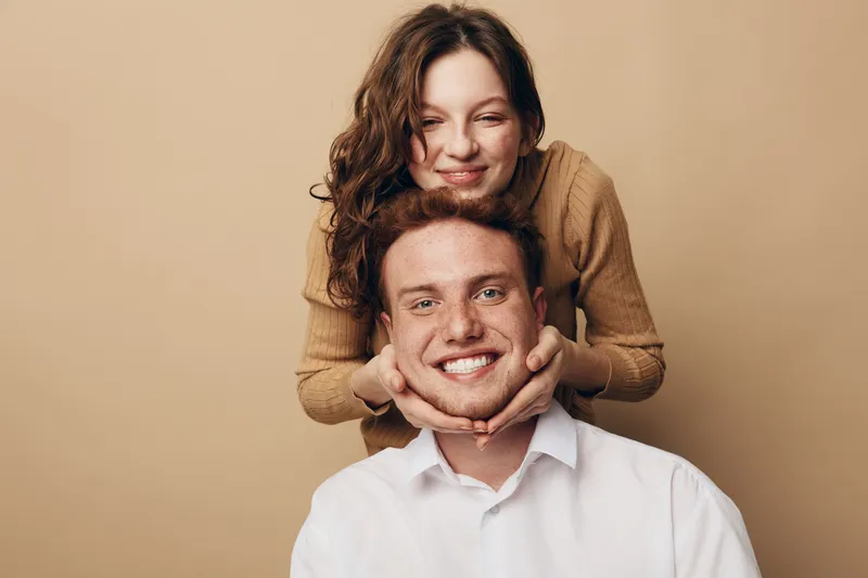 Smiling young woman with curly hair gently holds the face of a happy man with red hair and freckles. Warm beige background creates cozy and affectionate mood of friendship and love.
