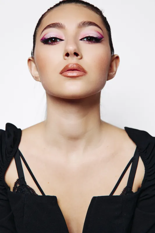 Young woman with artistic pink and glitter eye makeup looks confidently at camera. Beauty creative fashion portrait with smooth skin and sleek hair. Studio shot on white background.
