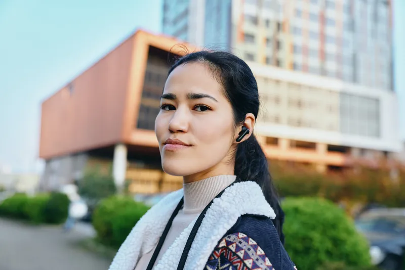 Woman with headphones in an urban street scene, outdoor city backdrop featuring modern buildings, stylish jacket and scarf, confident mood and casual fashion for lifestyle and travel visuals