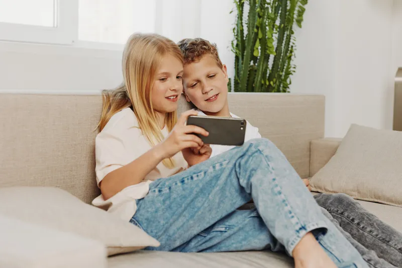 Two children sitting on a couch looking at a smartphone together, sharing a moment of joy and curiosity in a bright, modern living room setting with casual clothing.