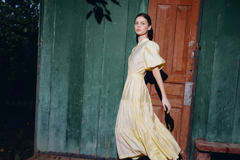 Fashionable woman in a flowing yellow dress walks past a weathered doorway in an outdoor setting, capturing a candid street style moment with soft evening light.