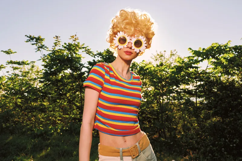 Vibrant woman outdoors in a bright striped top and sunflower sunglasses, sunny moment with a playful retro mood, lush greenery background, relaxed lifestyle vibe and natural colors