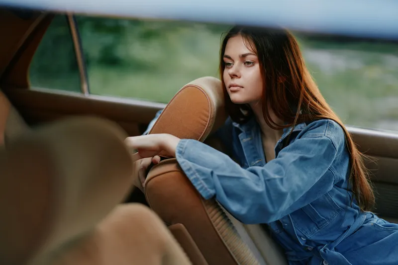 Thoughtful young woman in denim jacket resting in a vintage car with a green background, conveying emotions of nostalgia and contemplation.