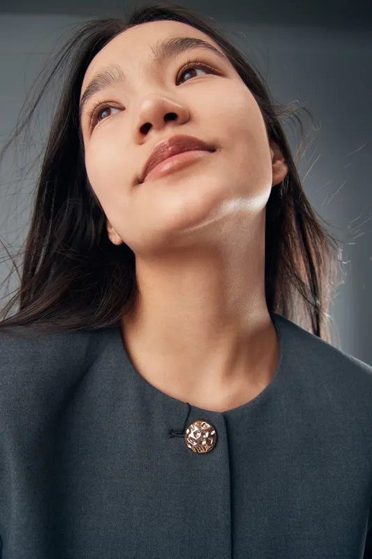Young Asian woman portrait looking upward with natural expression wearing dark jacket and button, studio lighting with neutral background and smooth skin detail closeup.