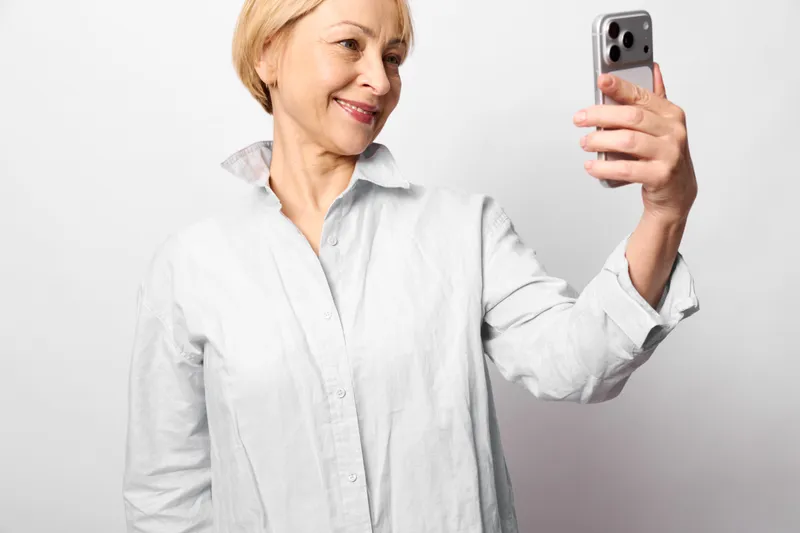 Smiling elderly woman takes a selfie with smartphone in hand, casual white shirt portrait on neutral white background, communication lifestyle technology mood