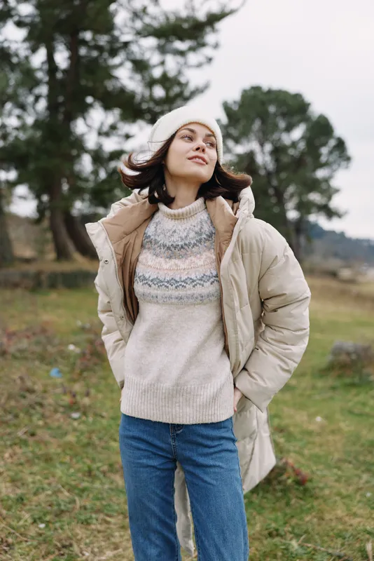 Woman standing in a light coat and knitted sweater in a field with trees exuding confident casual style and calm outdoor mood under soft daylight