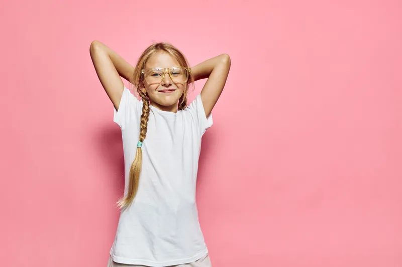 Smiling girl with glasses and braided hair stands relaxed with hands behind head on pink background.
