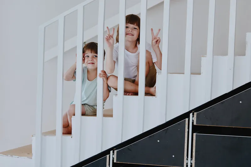 Two playful children happily posing on a staircase, showcasing joy and innocence with peace signs. Bright and modern interior setting enhances the mood.