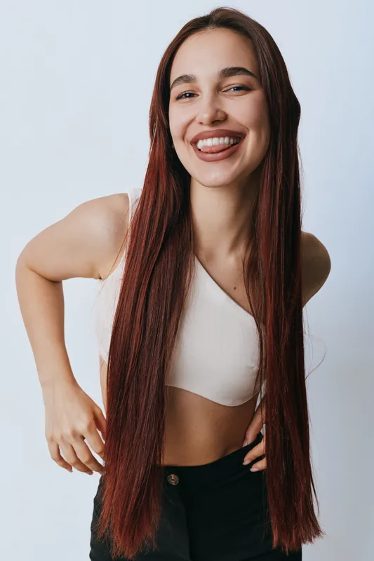 Young woman with long straight hair smiling and sticking out tongue wearing casual white top and black pants against white background in studio portrait.