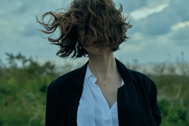 Young adult woman in a white shirt and black jacket stands in a windy outdoor field, conveying casual fashion and stylish travel vibe with moody atmosphere and confident pose for editorial
