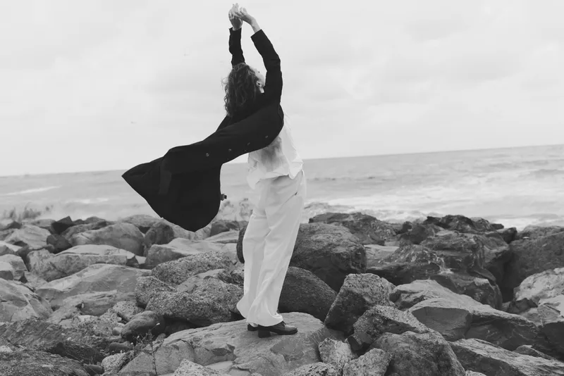 Dancing couple on a rocky coast, capturing movement and wind as they twirl, with ocean spray and monochrome tones, conveying drama, balance and freedom in an outdoor coastal scene