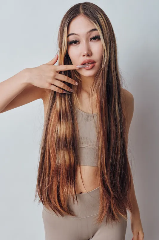 Young woman with long straight hair posing with hand near face wearing beige top and pants against plain white background in studio.