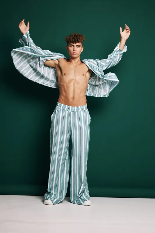 Young shirtless man in striped open jacket and loose pants posing with raised arms against dark plain background in studio. Fashion, style and confident male model expressing energy.
