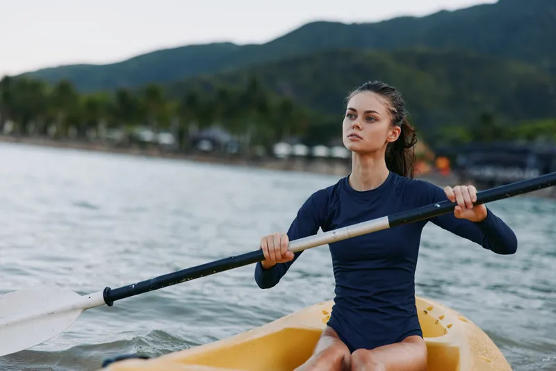 A focused female kayaker paddles a yellow kayak across calm water, gripping the paddle with determination as a scenic mountain backdrop frames the scene.