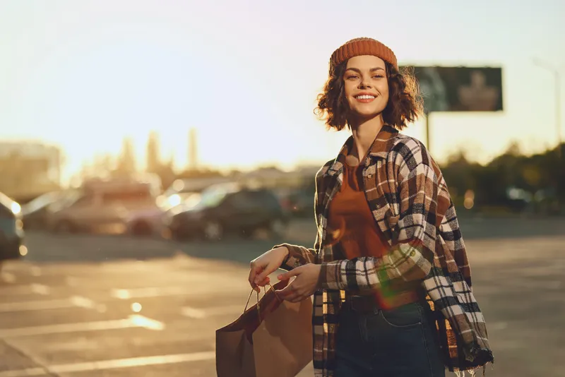 Woman shopping portrait with smile in golden hour, candid lifestyle scene in urban parking lot with paperbag and beanie, authenticity and golden hour glow evoke mindful living and emotional