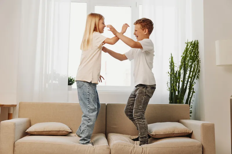 Two playful children jumping on sofa in bright living room, enjoying fun and energetic moment together at home.