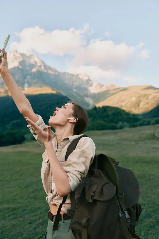 Young woman with backpack hiking in mountains holding smartphone up, enjoying nature and adventure, lifestyle concept with clear sky and scenic landscape background.