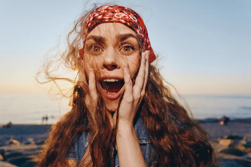 Lifestyle woman in modern boho-western denim street style with red bandana, surprised expression, film color tone, outdoor beach background, natural light.