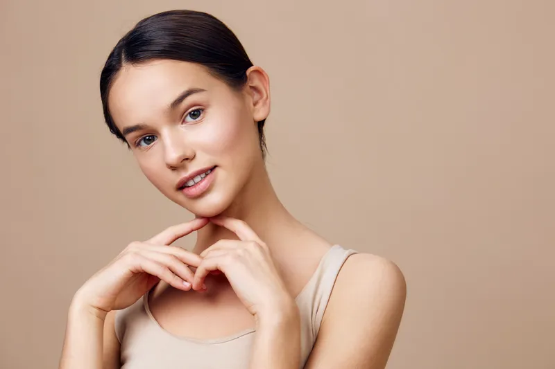 Young woman with natural beauty, wearing a beige top, smiling gently with a soft expression against a warm, neutral background, showcasing a healthy, minimalist style