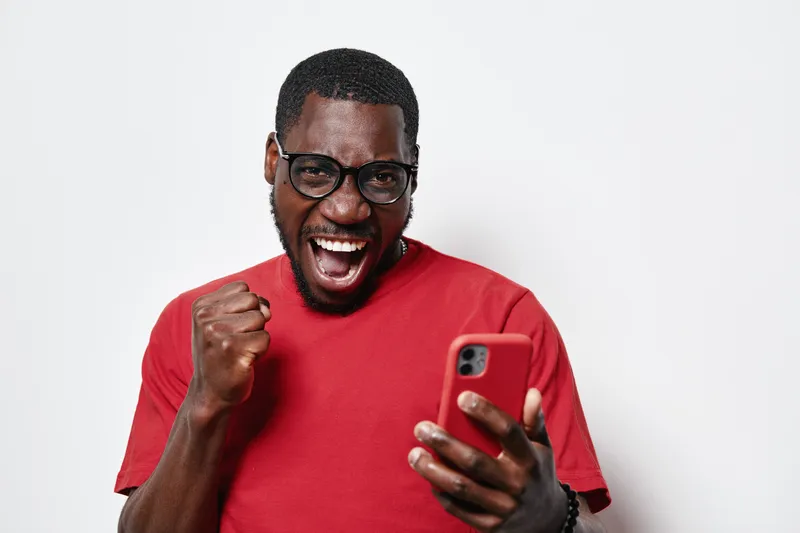 African American man with dark skin wearing glasses and red shirt holding smartphone celebrating success with fist pump and excited expression on white background.