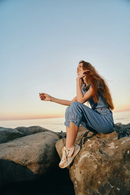lifestyle woman in modern boho-western denim street style sitting on rocks by the sea during sunset with warm film color tones and relaxed mood