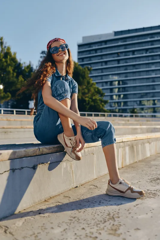 Lifestyle woman in modern boho-western denim street style wearing sunglasses and sneakers, sitting outdoors with urban building background, film color tone.