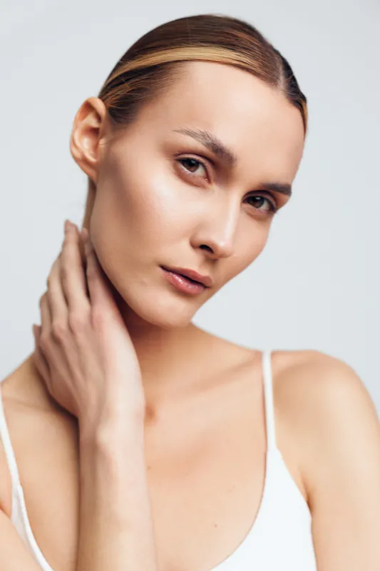 Young woman with clear skin posing with hand on neck against white background. Concept of natural beauty, skincare, and minimalism. Soft lighting highlights smooth complexion.