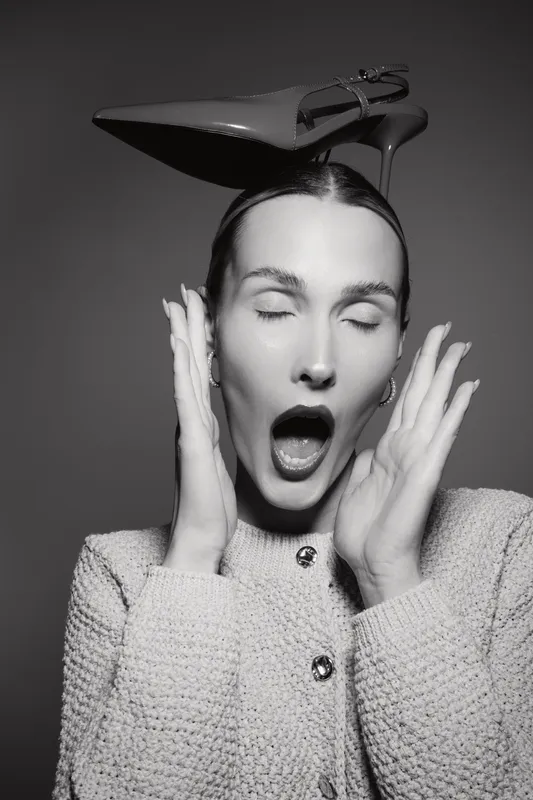 Caucasian woman with light skin wearing textured jacket and high heel shoe on head, eyes closed and mouth open in studio. Concept for fashion and expression.