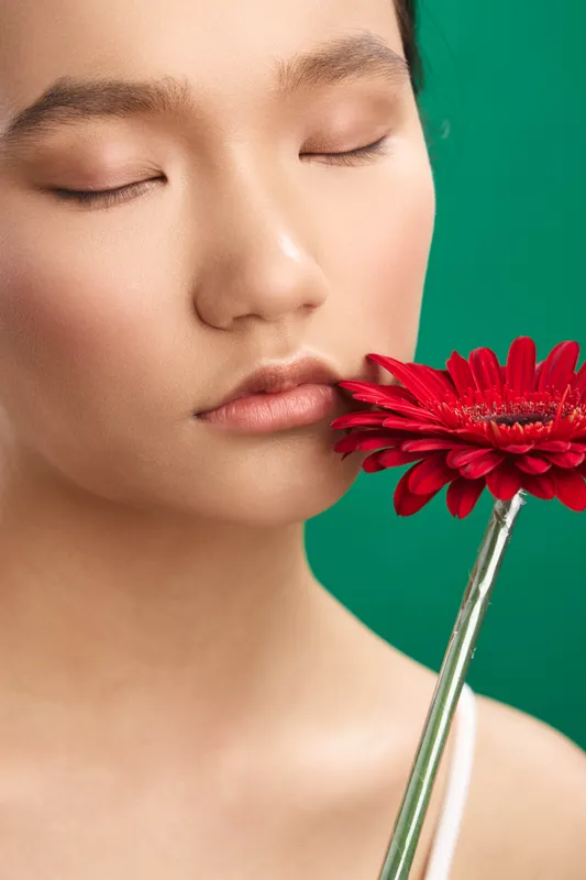 Asian woman with serene expression holds a red flower near her lips in a close up portrait. Soft lighting and a vibrant green backdrop create an intimate beauty moment.
