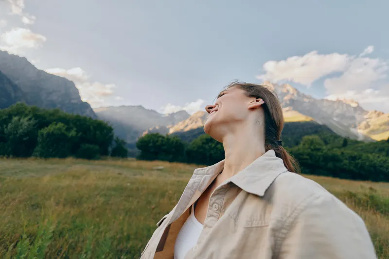 Young woman in a beige jacket gazes up at the sky, standing in a wide grass field with rugged mountains in the background, enjoying soft natural light and a sense of freedom.