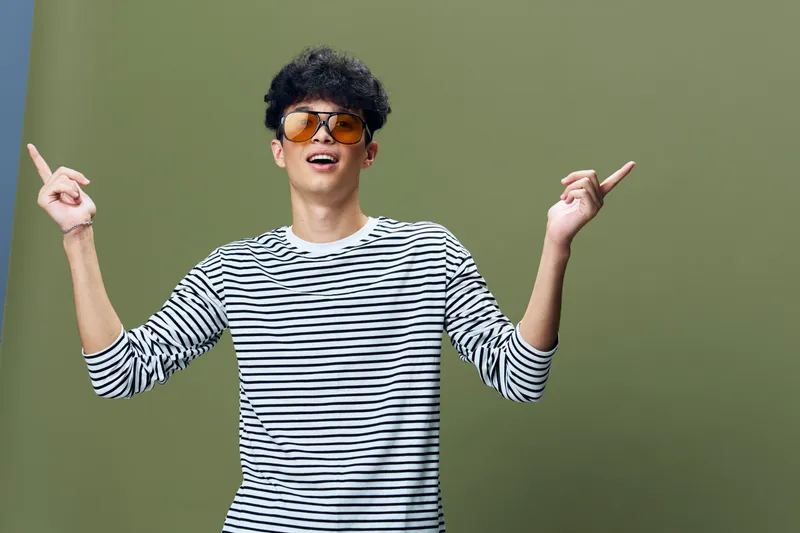 Young man wearing striped shirt and sunglasses smiling and pointing up with both hands against green background, casual style, positive mood.