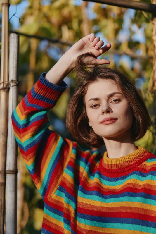 Woman portrait in rainbow sweater outdoors with warm sunlight and autumn leaves background, colorful knit closeup showing short bob hairstyle, natural beauty and confident casual fashion pose.