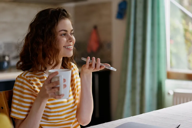 A smiling woman sits at a home kitchen table, striped shirt, holding a mug and a phone. A cozy morning routine concept for lifestyle, comfort, and relaxed domestic living.