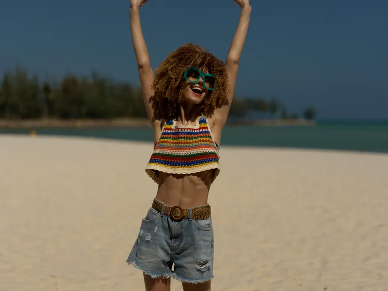 Smiling woman with curly hair wearing colorful top and denim shorts enjoying sunny beach day with sunglasses and raised arms, expressing happiness and freedom.