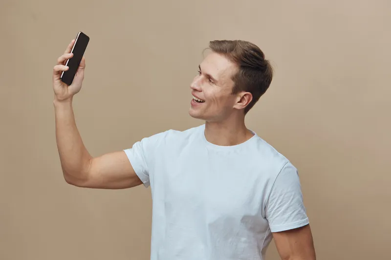 Smiling young man taking a selfie with smartphone, casual clothing, cheerful mood, beige background, studio portrait, lifestyle concept, indoor.