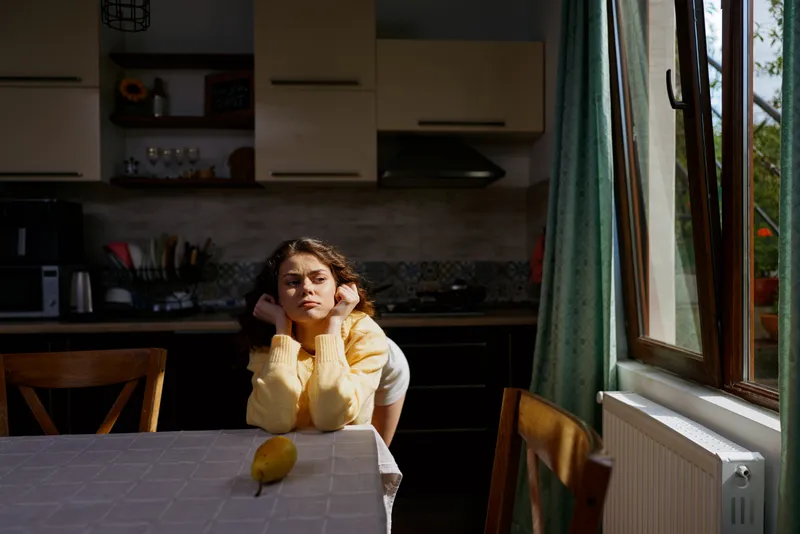 Young woman sits alone at a kitchen table, soft natural light from a window, pensive mood, home lifestyle scene, everyday moment indoors, quiet domestic atmosphere.