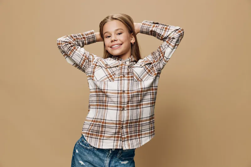 Girl smiling with hands behind head wearing checked shirt and ripped jeans standing against beige background with relaxed and confident pose in studio.