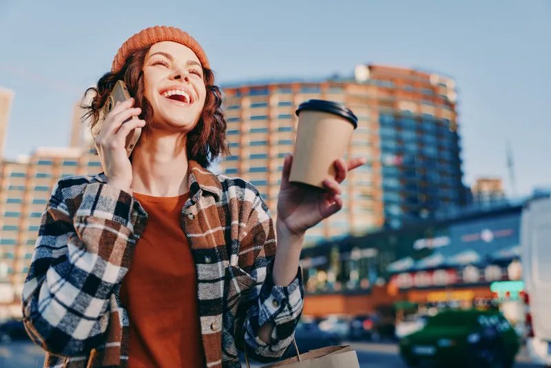 woman coffee phone city smile urban laughing beanie young woman talking on mobile while holding takeaway cup, golden hour glow and candid lifestyle mood with authenticity and emotional storytelling.