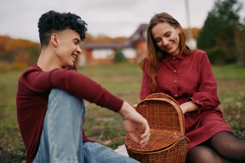Two friends share an authentic moment outdoors, sitting on grass with a woven basket, honest smiles, and relaxed posture, conveying truth through simple connection.