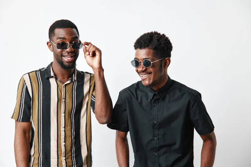 Two young African American men with dark skin wearing sunglasses smiling and standing together in casual striped and black shirts on white background.
