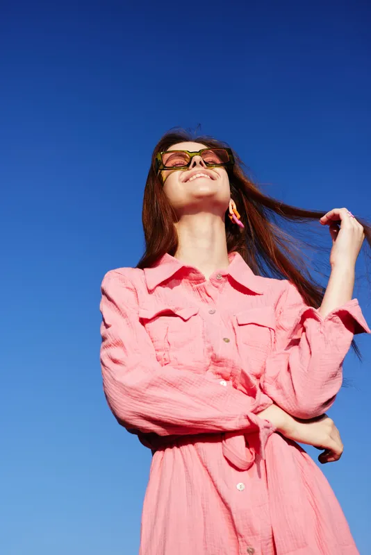 Smiling young model in a pink dress enjoying a sunny day against a clear blue sky, conveying joy and positivity, perfect for lifestyle and fashion themes