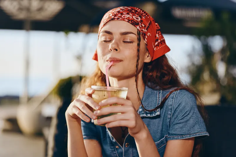 Lifestyle woman enjoying drink in modern boho-western denim street style with red bandana and natural sunlight, captured in warm film color tone.
