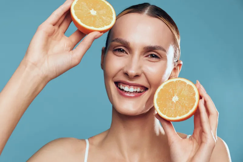 Woman holding orange slices, citrus smile, skincare on face, studio portrait, blue background, fresh fruit, soft light, beauty glow, clean healthy skin, hydration