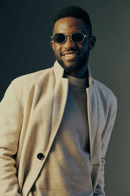Stylish young Black man wearing sunglasses and a beige coat, smiling confidently against a muted background in a modern fashion context