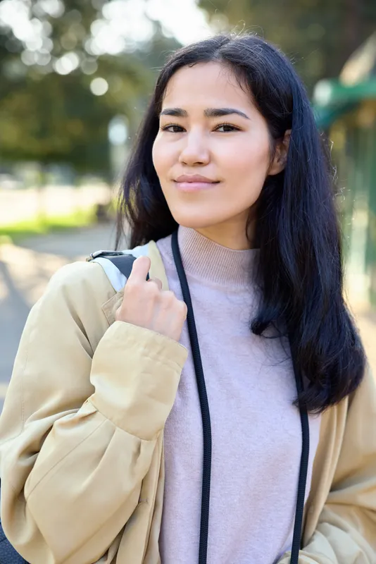 Woman in a light sweater and beige cardigan stands outdoors in a sunlit park with a backpack strap over her shoulder, conveying casual travel and outdoor style.